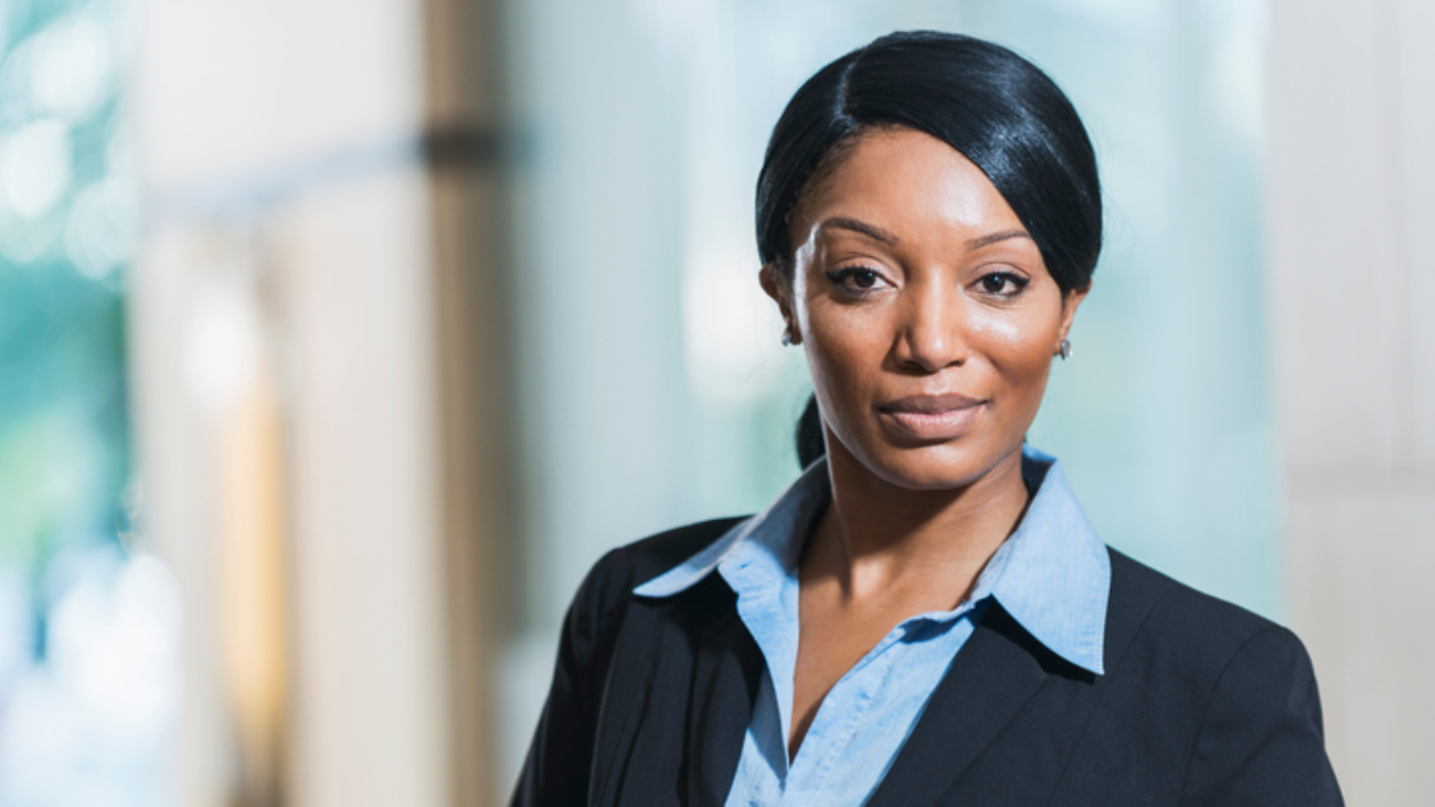 Portrait of an African American businesswoman standing in an office lobby, near a window, looking at the camera with a serious, confident expression.  She has straight, long, black hair hair back in a ponytail.  She is wearing a black suit.